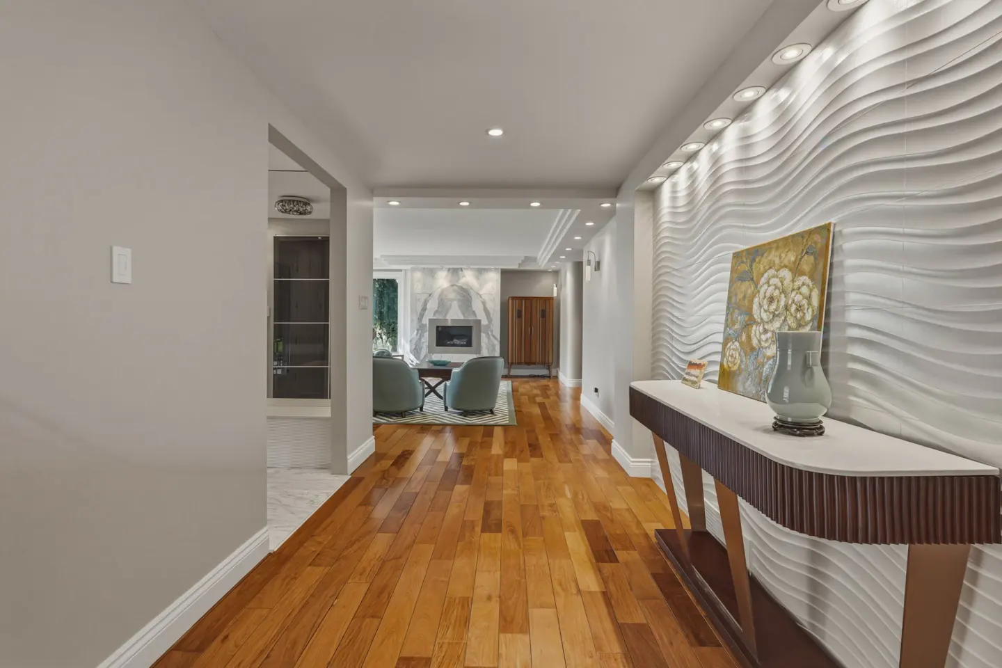 Hallway with hardwood floors, a textured white wave wall, a console table with art, and a view into a modern living room.