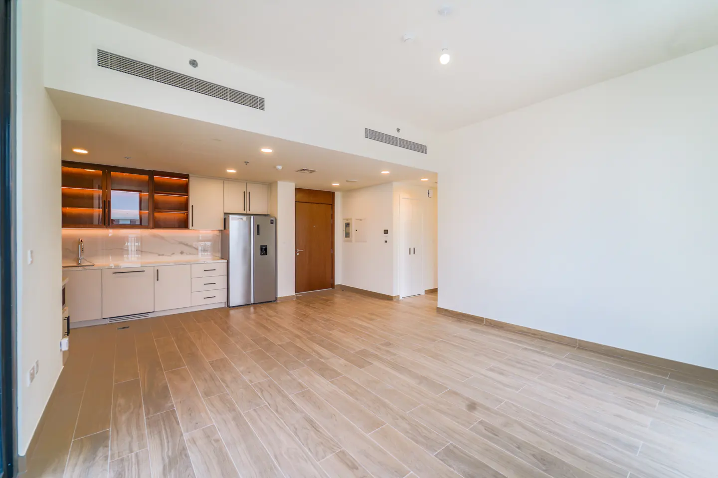Bright, empty apartment with wood-look tile floors, white walls, and a kitchen with stainless steel refrigerator.