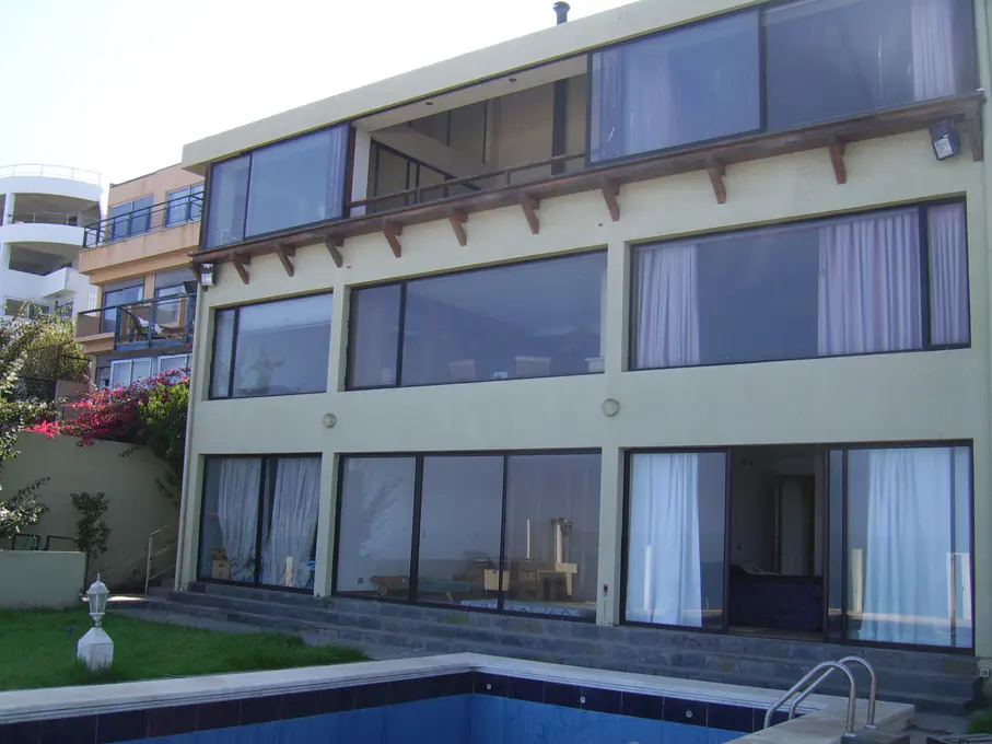 Three-story beige house with large windows, a pool, and a green lawn. Other buildings are visible in the background.