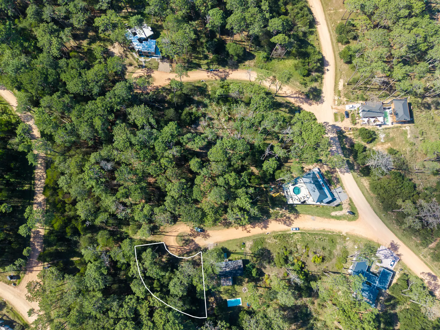 Aerial view of a wooded lot outlined in white, surrounded by green trees and dirt roads. Houses are visible in the background.