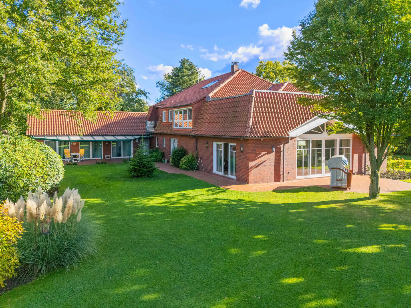 A red brick house with a red tile roof sits on a large green lawn under a blue sky.