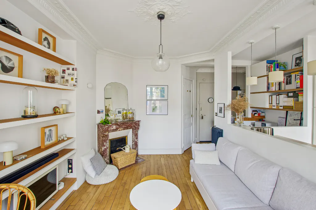 Bright living room with white walls, wood floors, and built-in shelves. A marble fireplace, white sofa, and round coffee table add charm.