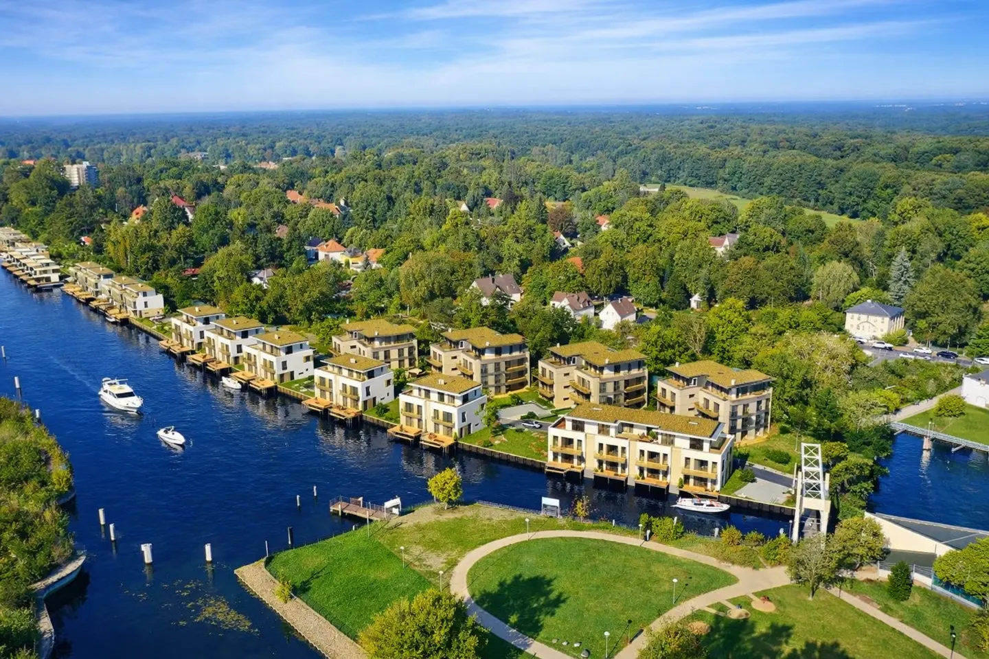Aerial view of modern waterfront homes along a blue canal, surrounded by lush green trees and a clear blue sky.