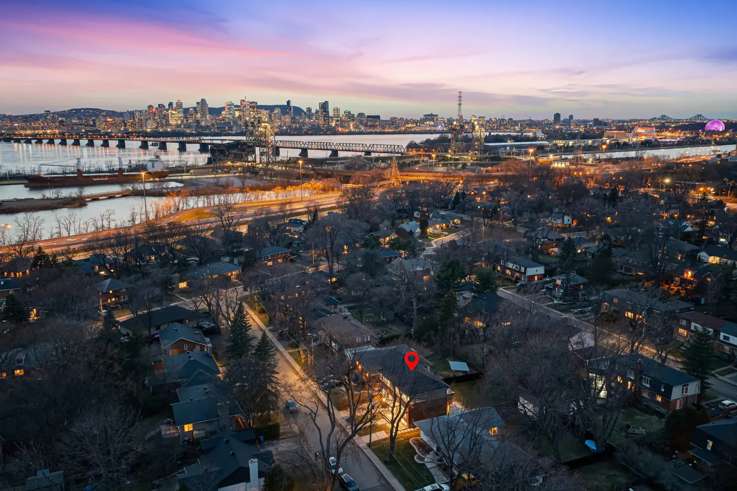 Aerial view of a house with a red pin on the roof, city skyline and river in the background at dusk.