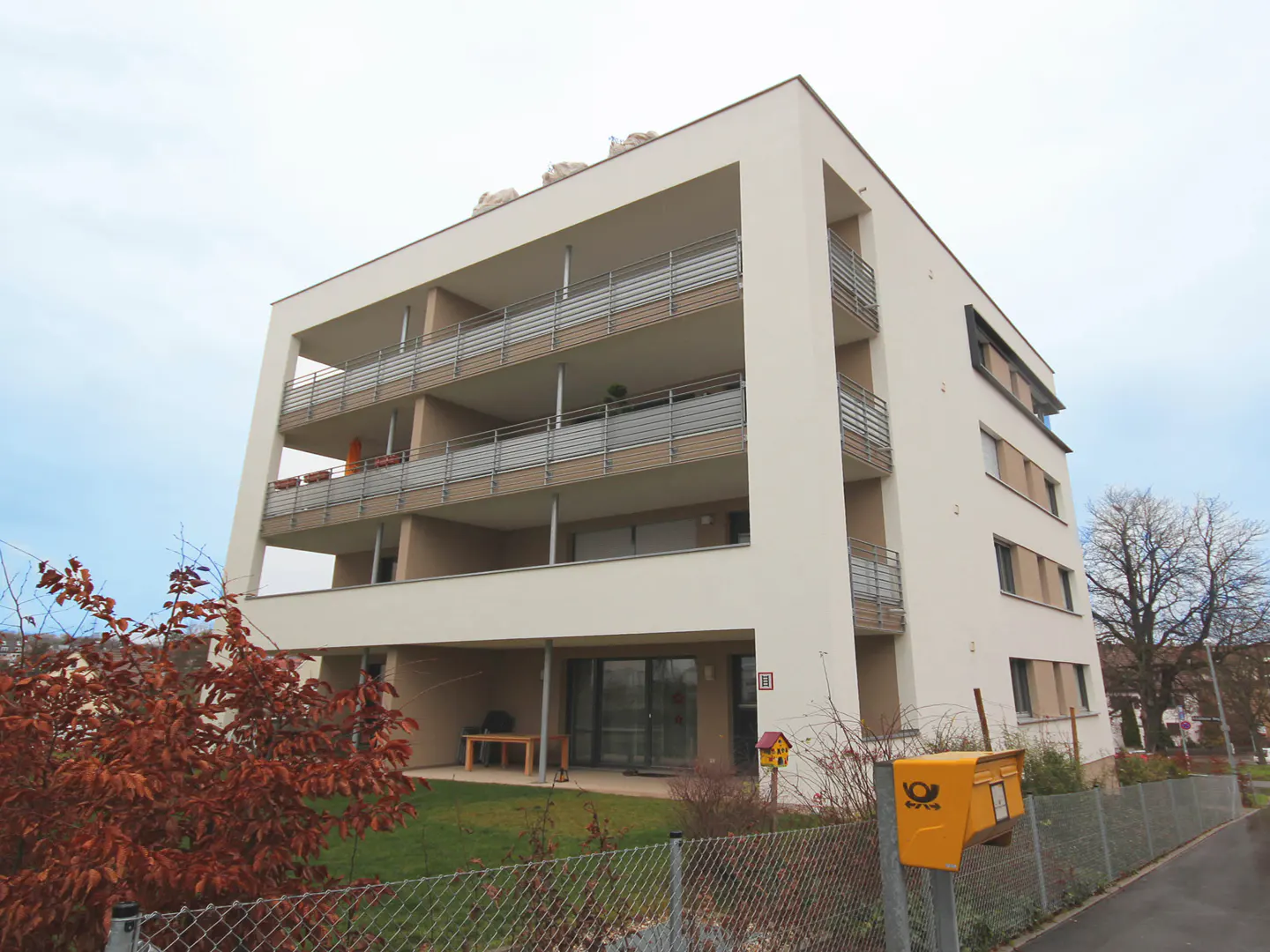 Modern, three-story apartment building with balconies, a yellow mailbox, and a chain-link fence.