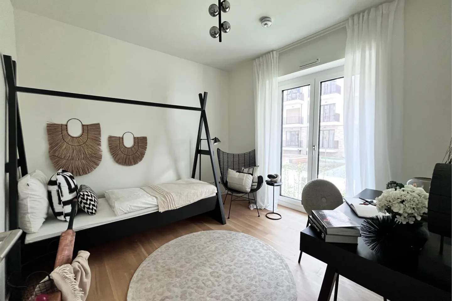 A modern bedroom with a black frame bed, white walls, and a round patterned rug. A desk with flowers sits near a window with white curtains.