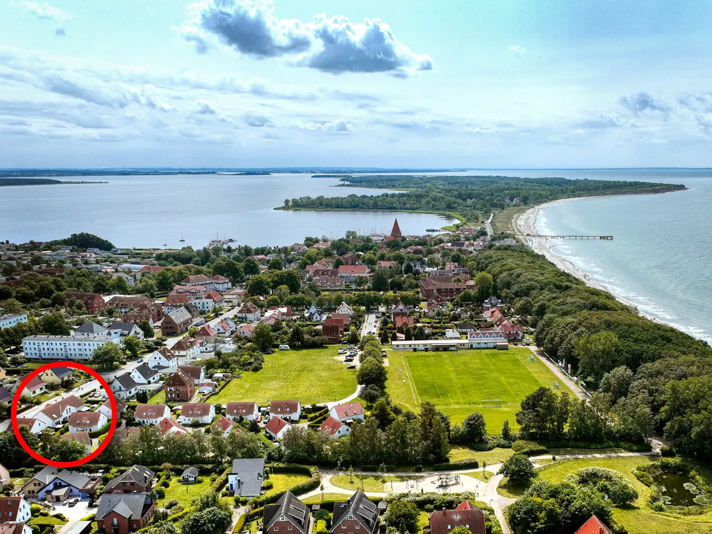 Aerial view of a coastal town with red-roofed houses, green fields, and a sandy beach, with a red circle highlighting a group of houses.