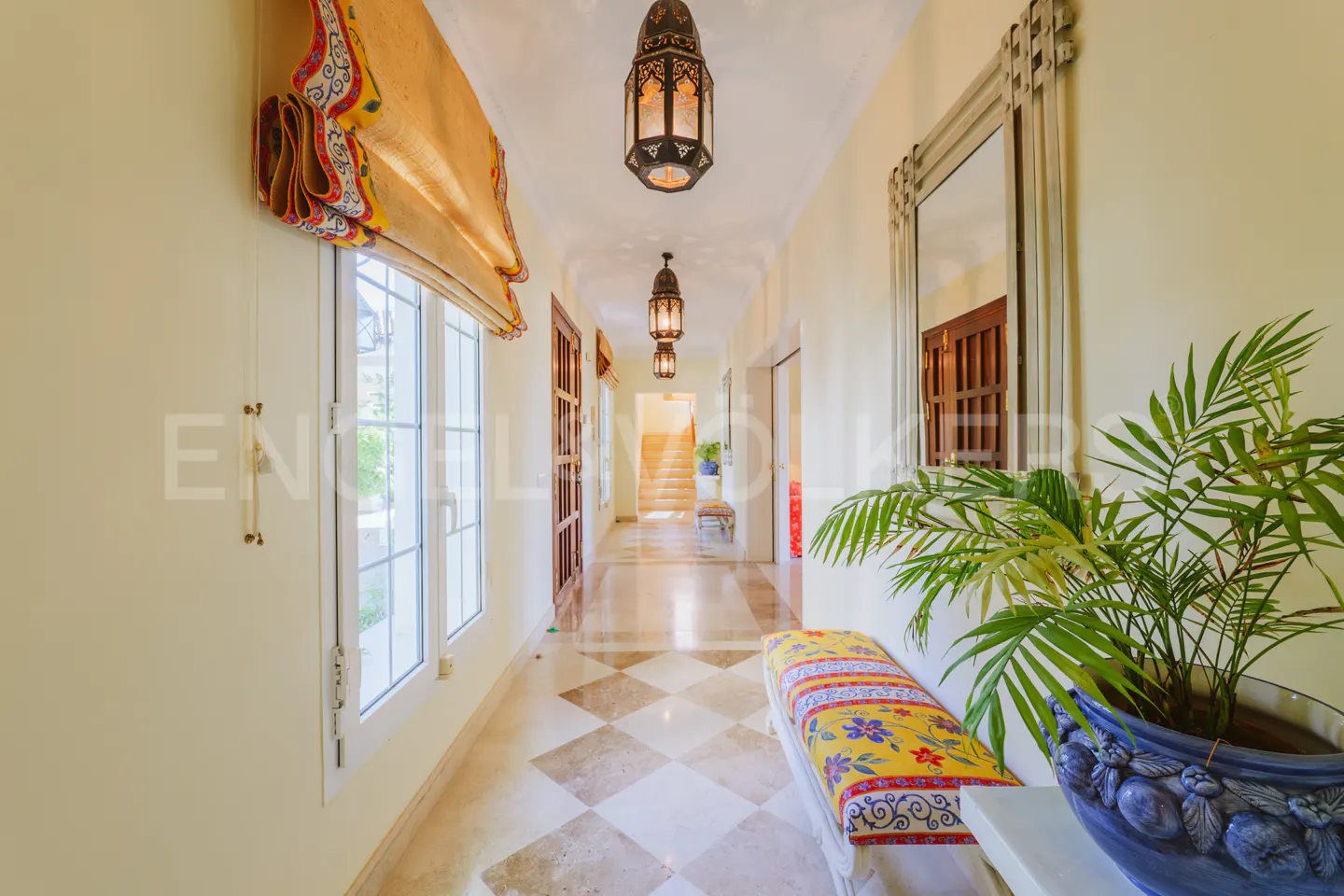 Hallway with cream walls, diamond-patterned floor, and hanging lanterns. A yellow floral bench sits near a potted plant. Windows with yellow shades line the left.