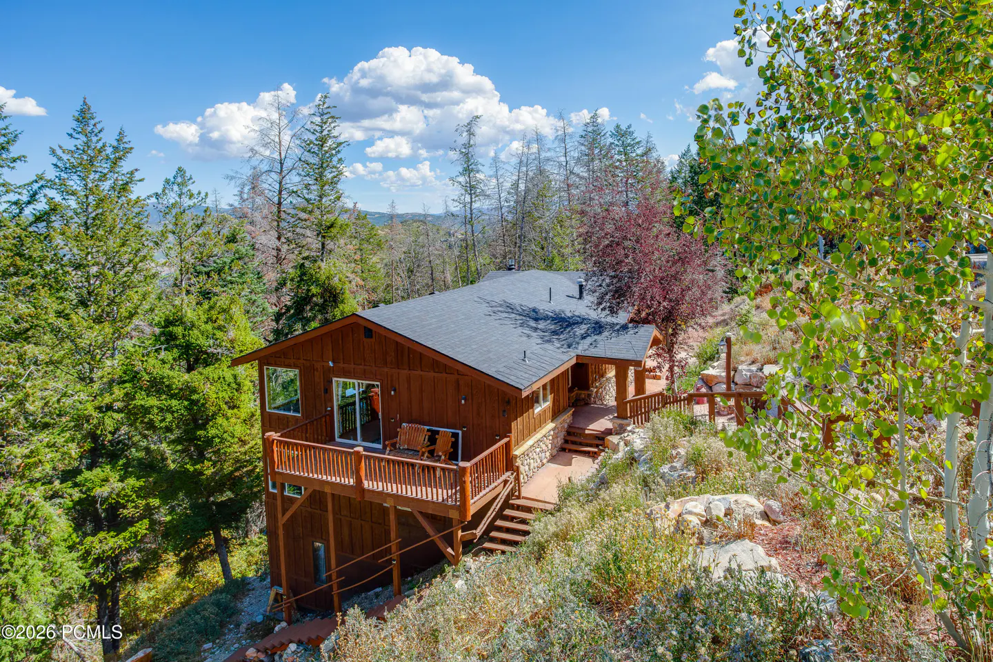 A brown house with a deck sits on a hill surrounded by trees under a blue sky with white clouds.