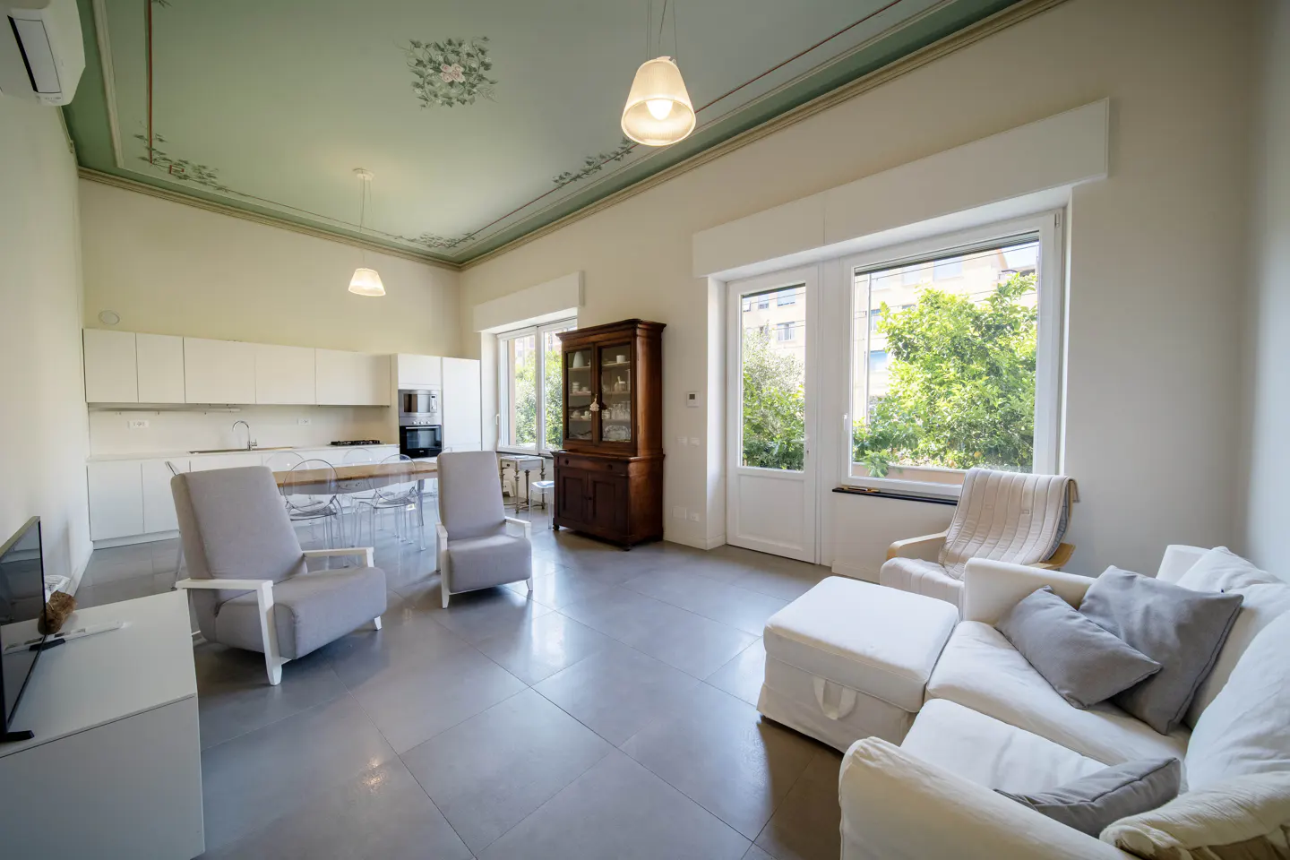 Bright, modern living space with white sofa, gray chairs, and kitchen. Large windows overlook green trees. Ornate ceiling detail.