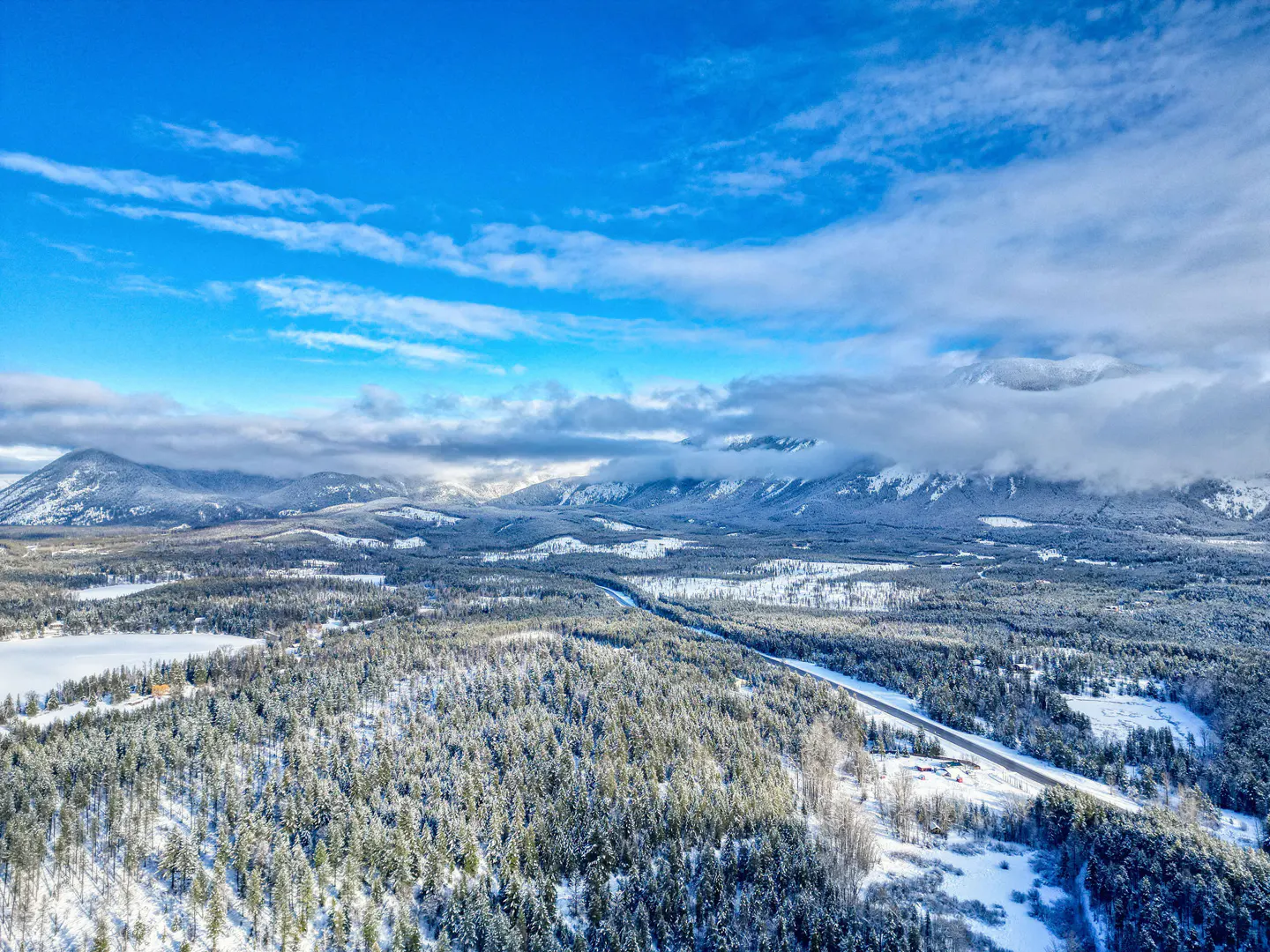 Aerial view of a snow-covered forest and mountains under a blue sky with white clouds. A road cuts through the trees.
