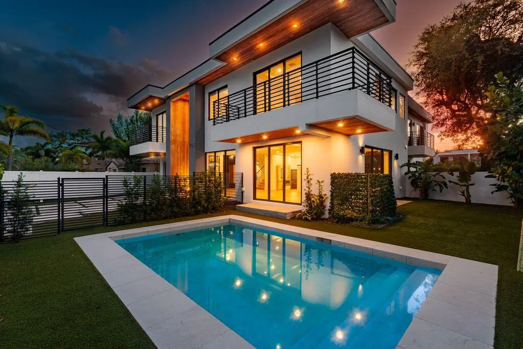 Modern two-story white house with a pool at dusk. Balconies have black railings. Wood accents and landscape lighting.