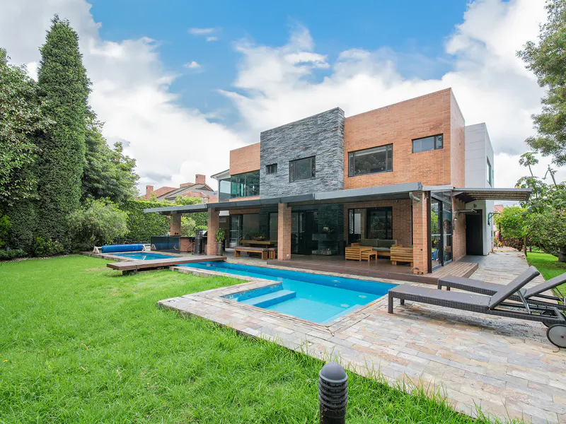 Backyard view of a modern brick house with a blue pool, lounge chairs, green grass, and trees under a cloudy sky.