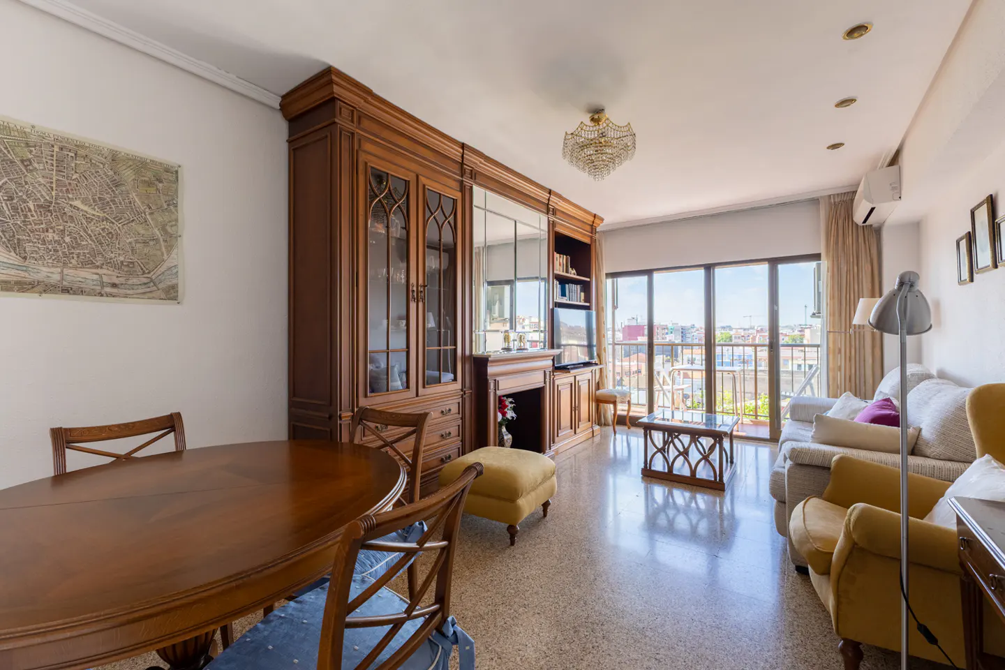Living room with a large wooden cabinet, dining table, and seating area. Balcony with city view in the background.