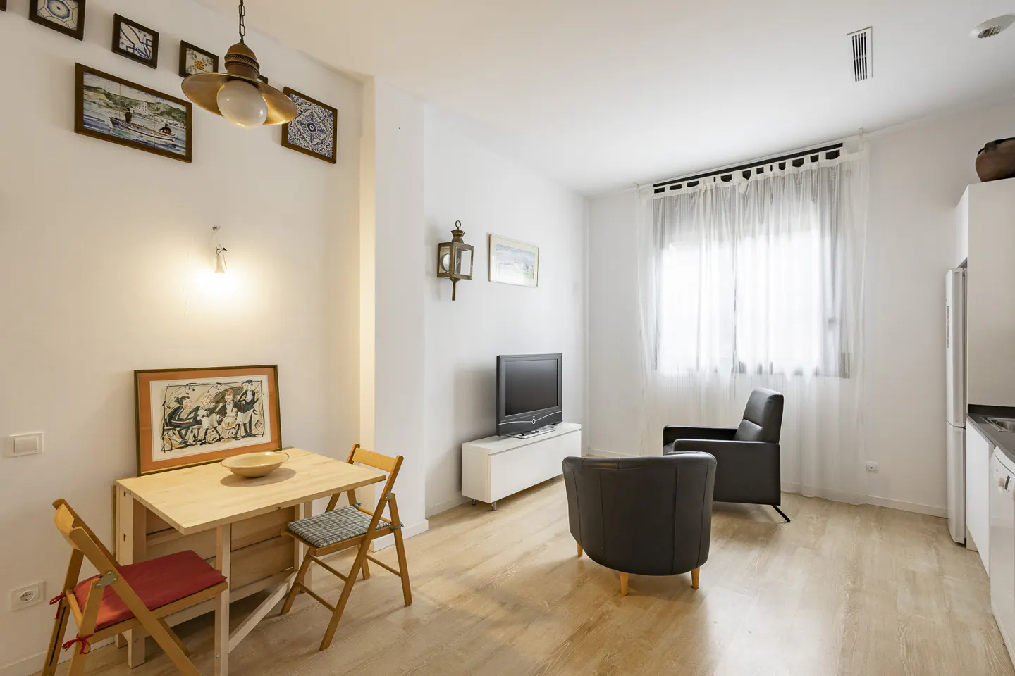 Bright living room with light wood floors, a small dining table, two dark armchairs, a TV, and white walls with art.