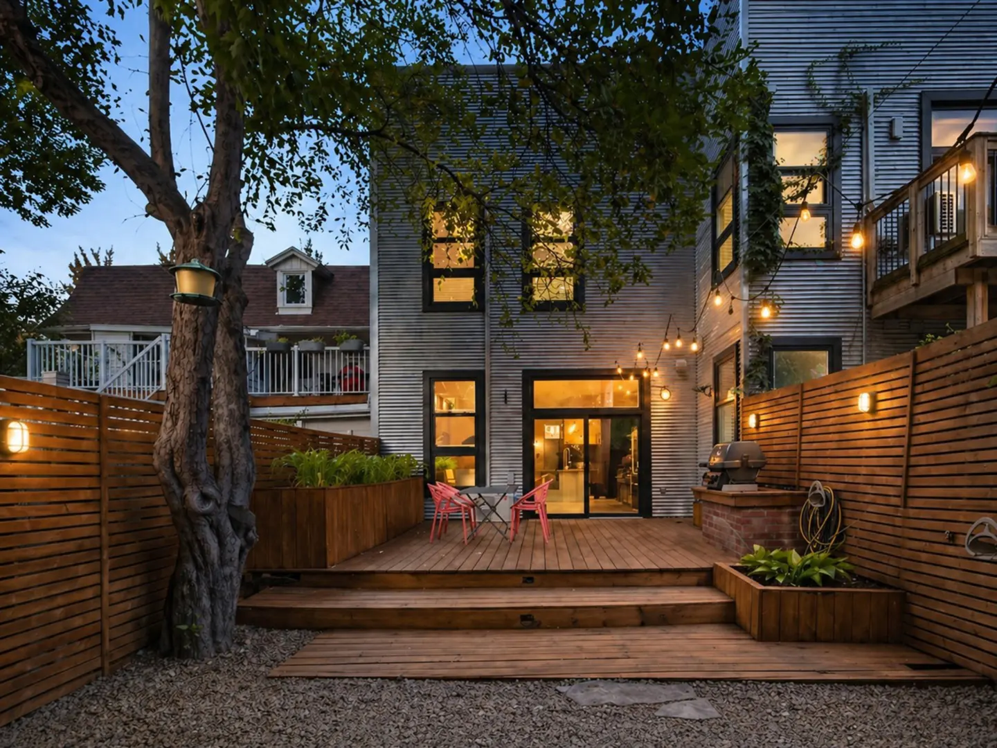 Backyard view of a two-story house with a wooden deck, fence, string lights, and a tree. Two pink chairs and a table are on the deck.