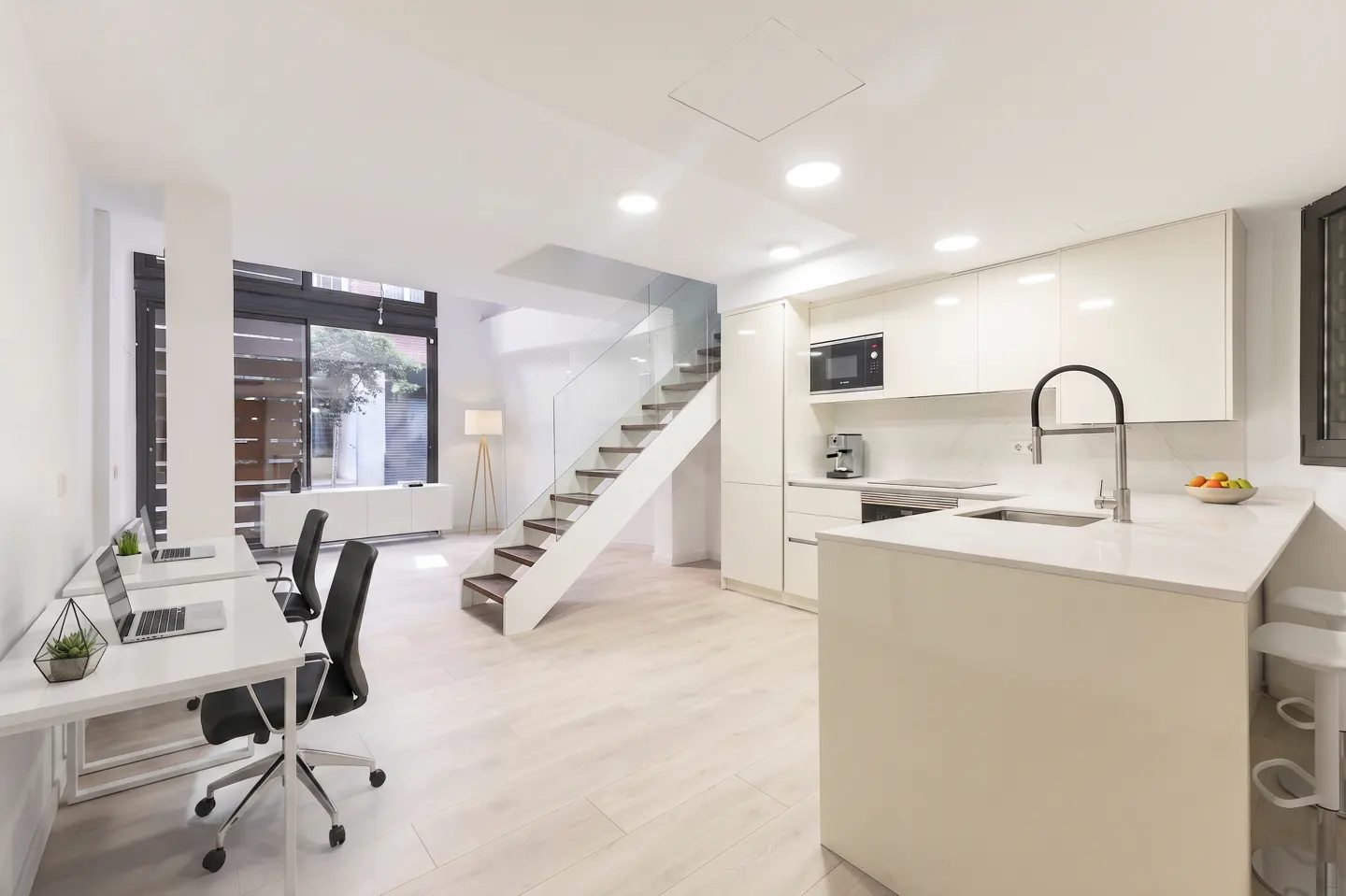 Bright, modern open-concept space with white walls, light wood floors, kitchen island, and office area with desks and laptops. A staircase leads to an upper level.