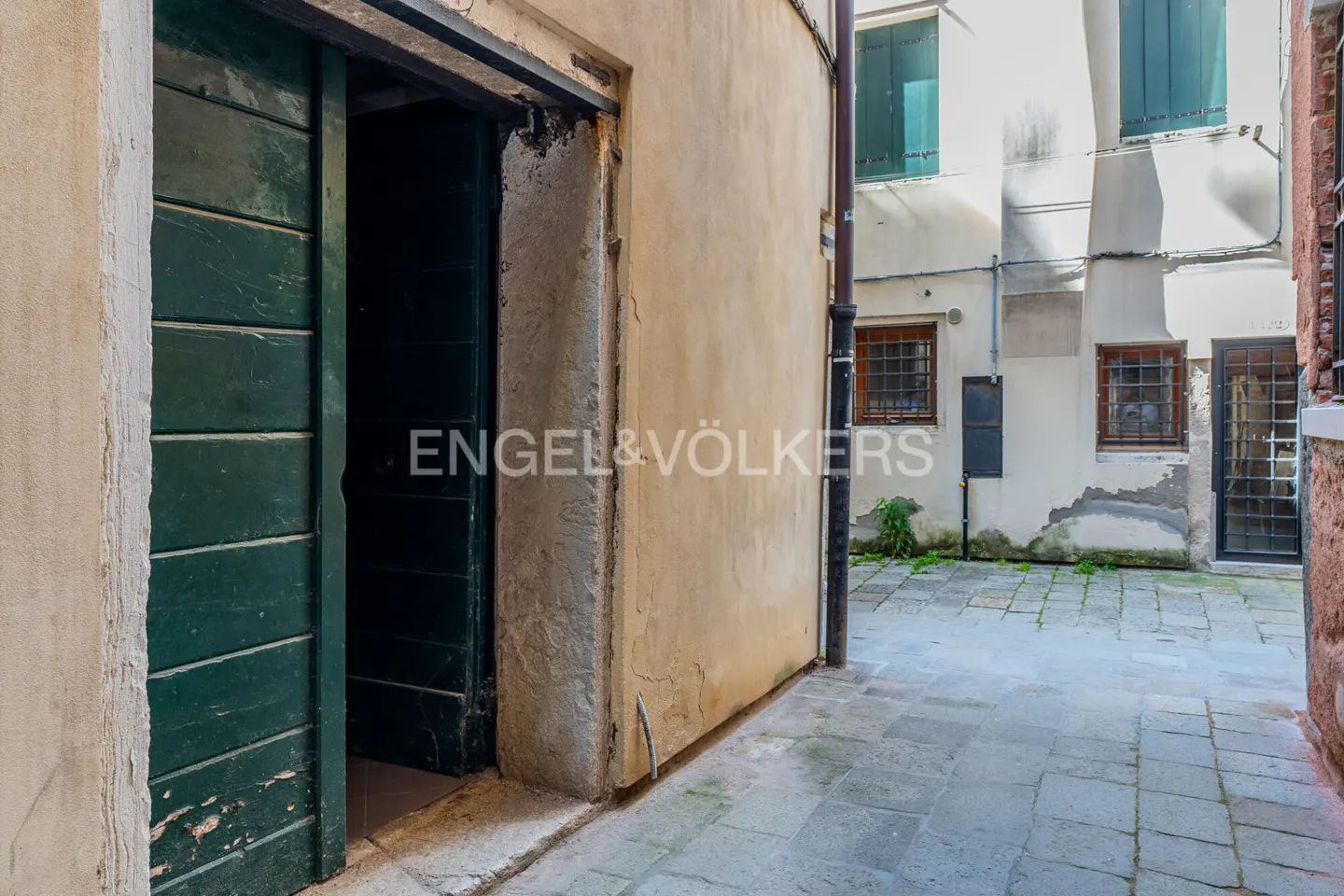 Exterior view of a narrow alleyway with a green door ajar, leading to a courtyard with buildings in the background.