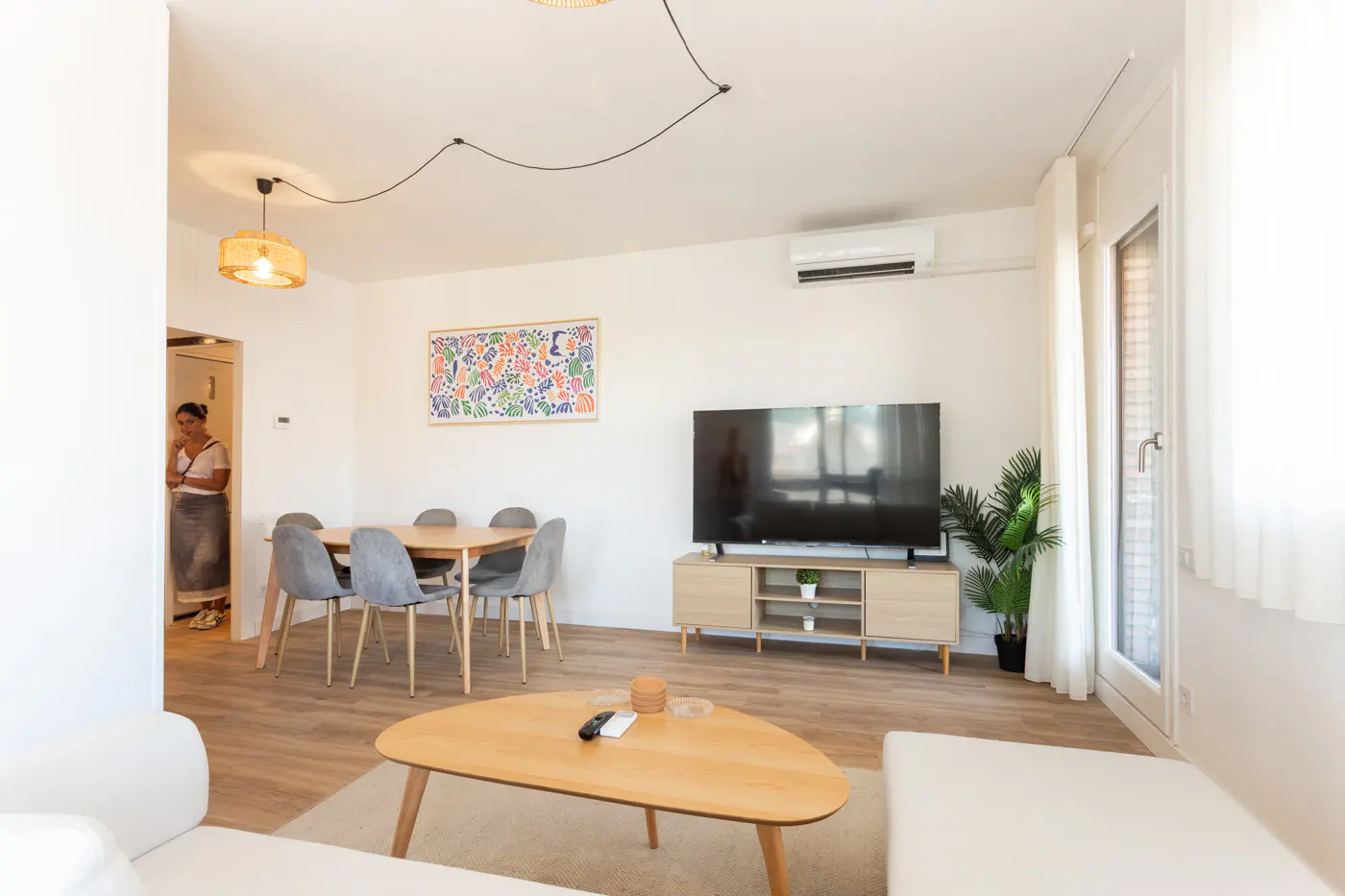 Bright living room with a wooden coffee table, TV on a media console, dining area with grey chairs, and a person in doorway.