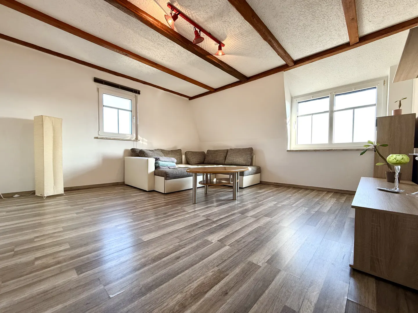 Attic living room with wood beams, gray wood floors, white walls, and a gray and white sectional sofa. Two windows provide natural light.
