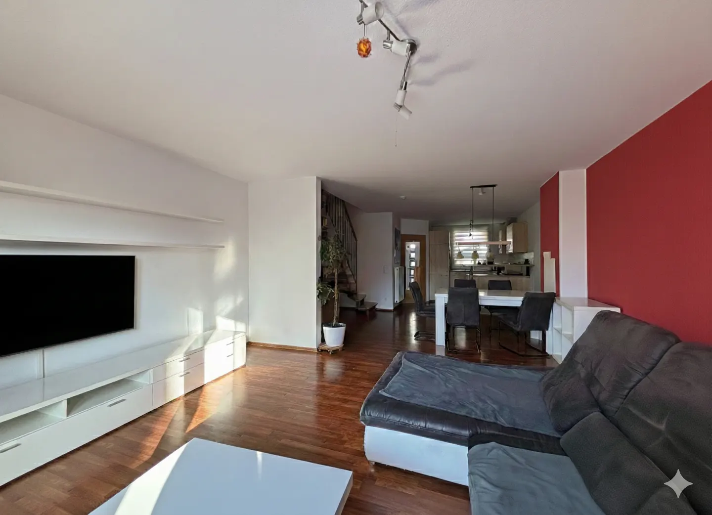 A modern living room with a gray couch, white TV stand, and a red accent wall. A dining table and kitchen are visible in the background.