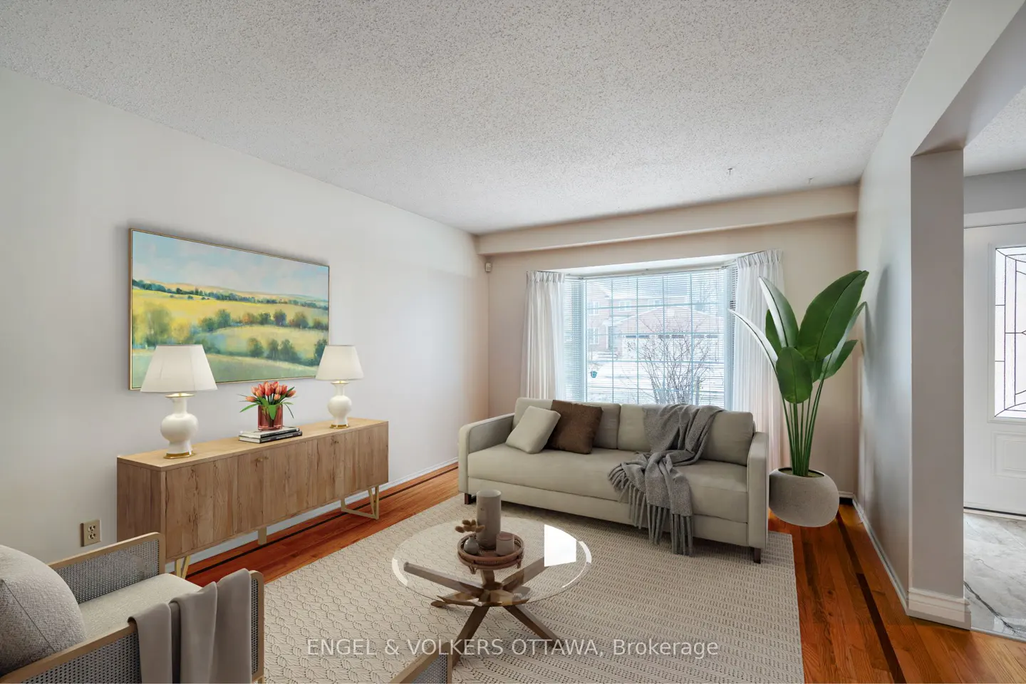 Living room with a beige sofa, glass coffee table, and light wood cabinet with lamps. A large window provides natural light.
