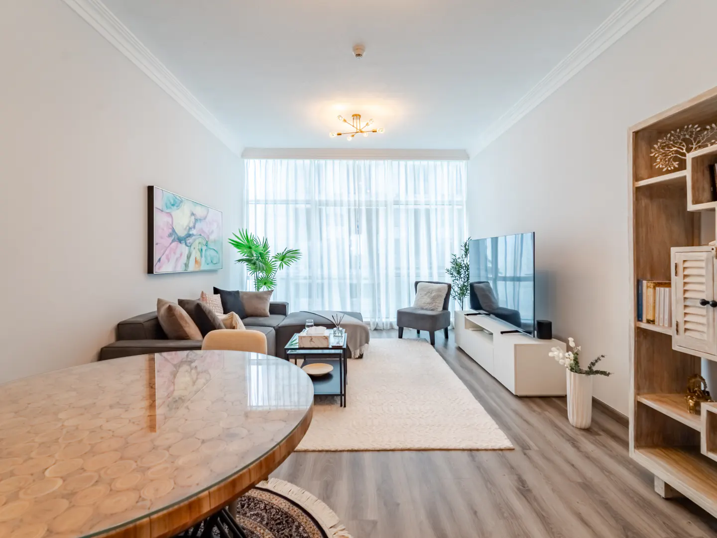 Bright living room with gray sectional, TV, and round wood table. White walls, sheer curtains, and light wood floors create an airy feel.