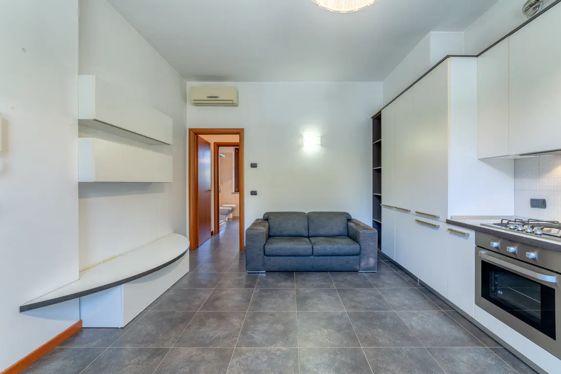 A modern apartment interior with white walls, gray tile floors, and a gray sofa. A kitchen area with white cabinets and stainless steel appliances is visible.
