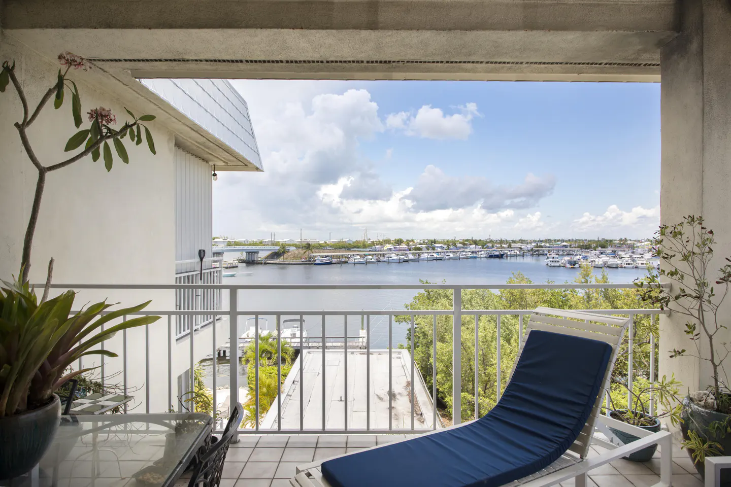 Balcony view of a marina. White railings, blue lounge chair, table, and potted plants. Water, boats, and sky in the background.