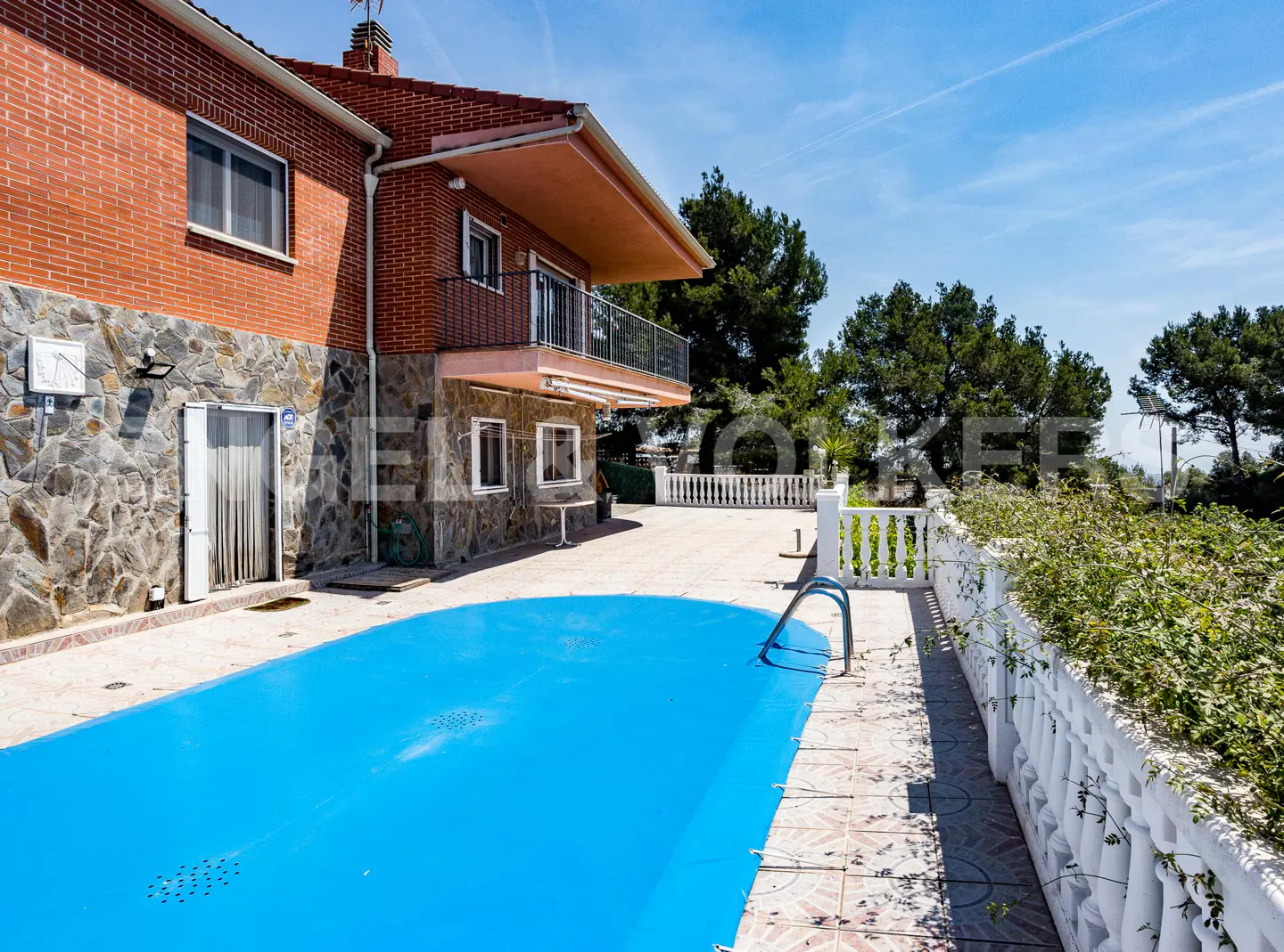 Exterior view of a brick and stone house with a blue covered pool and white fence.