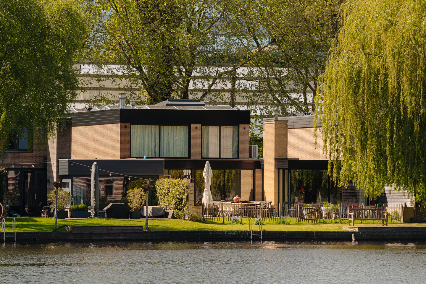 Modern waterfront home with brick and black trim, surrounded by green trees and lawn, reflecting in the water.