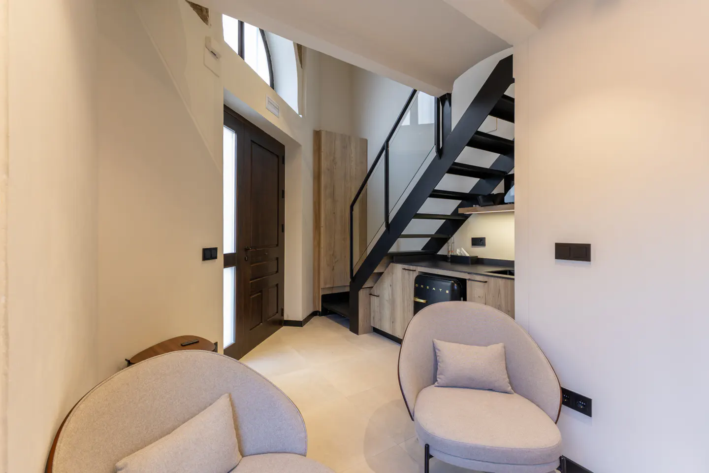 Interior view of a modern apartment with two gray chairs, a black staircase, and a small kitchen area.
