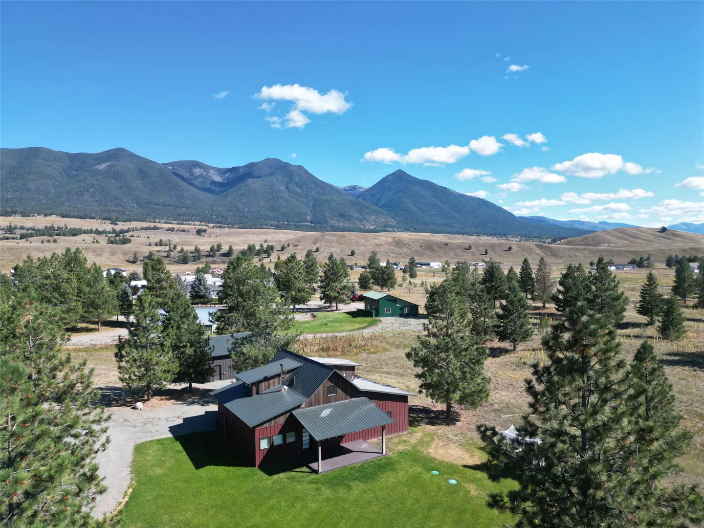 Aerial view of a red house with a green metal roof, surrounded by trees and mountains under a blue sky.