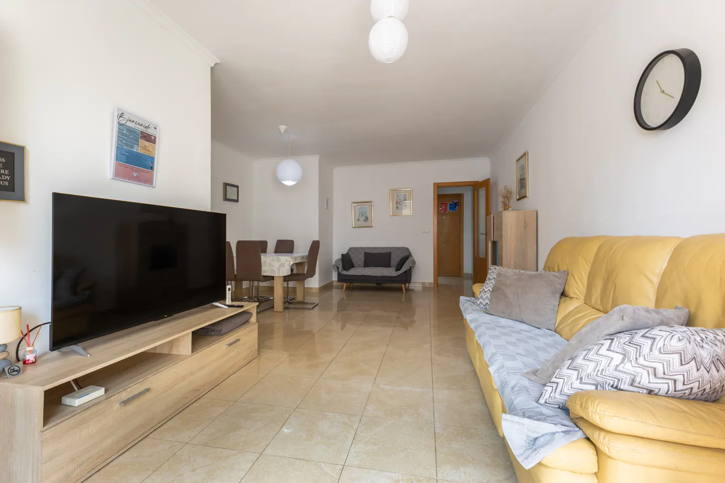 A bright living room with a yellow sofa, TV, dining table, and tiled floor.