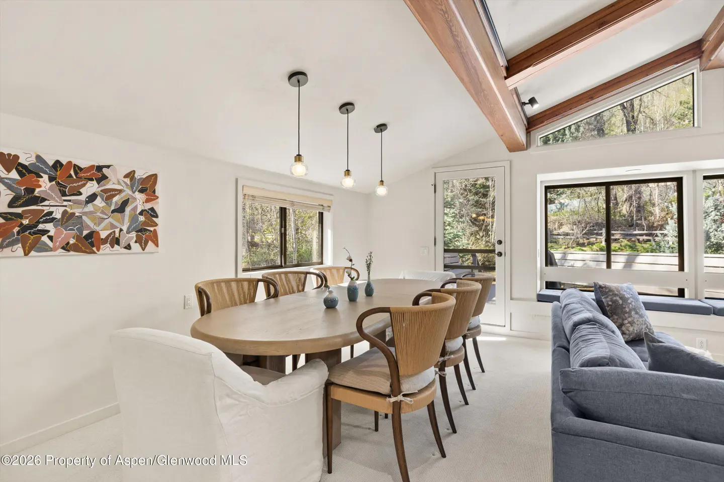 Bright dining room with an oval table, woven chairs, and modern pendant lights. Large windows offer views of the trees outside.