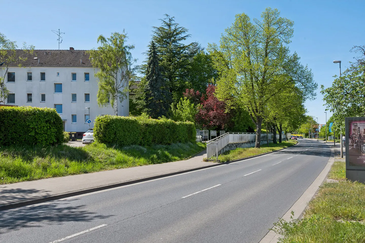 Street view of a gray asphalt road with white lines, green trees, and a white apartment building on a sunny day.