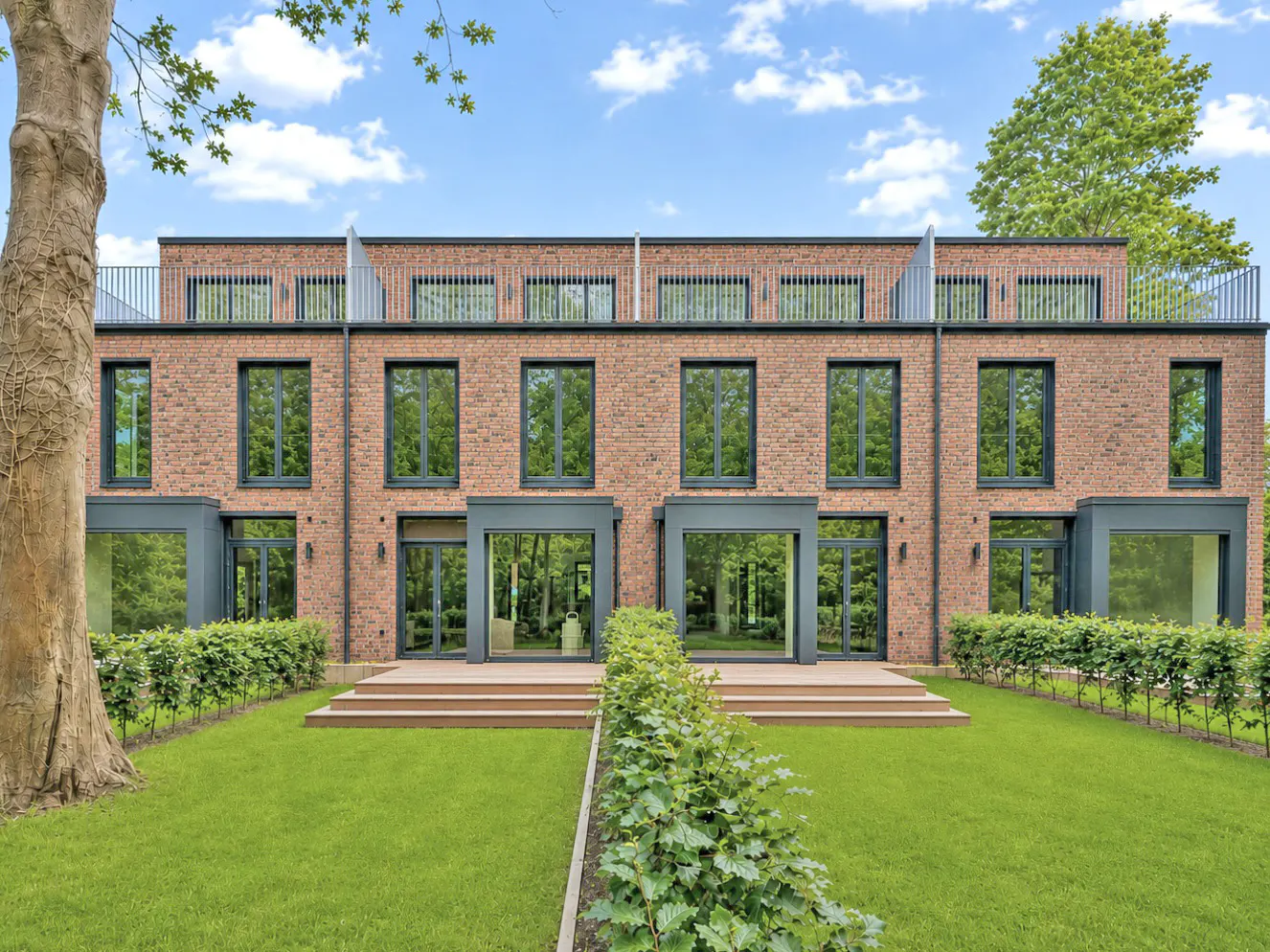 Modern red brick townhouse with large windows, a green lawn, and a wooden deck under a bright blue sky.