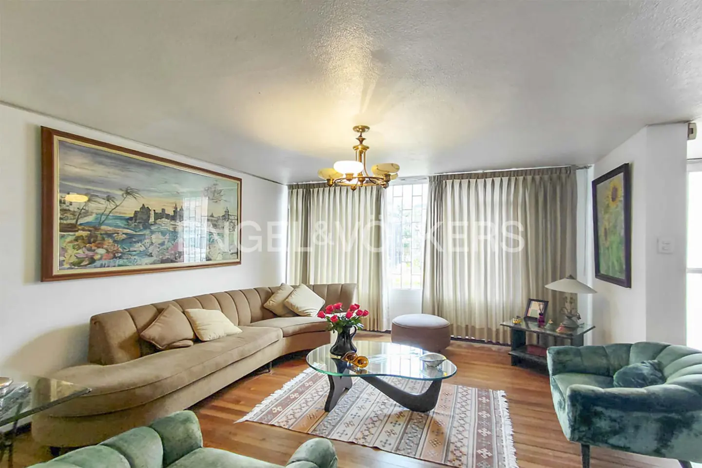 A living room with a brown sofa, green chairs, and a glass coffee table with red roses. A large painting hangs on the wall.