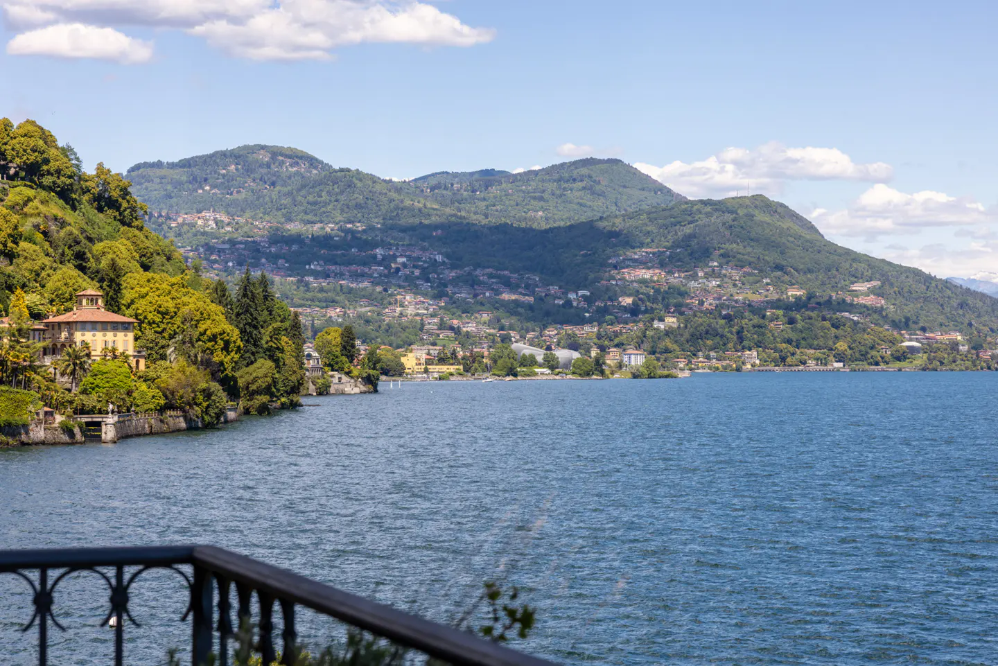 Lake view with blue water, green mountains, and a yellow villa nestled among trees on the shore. A black railing is in the foreground.