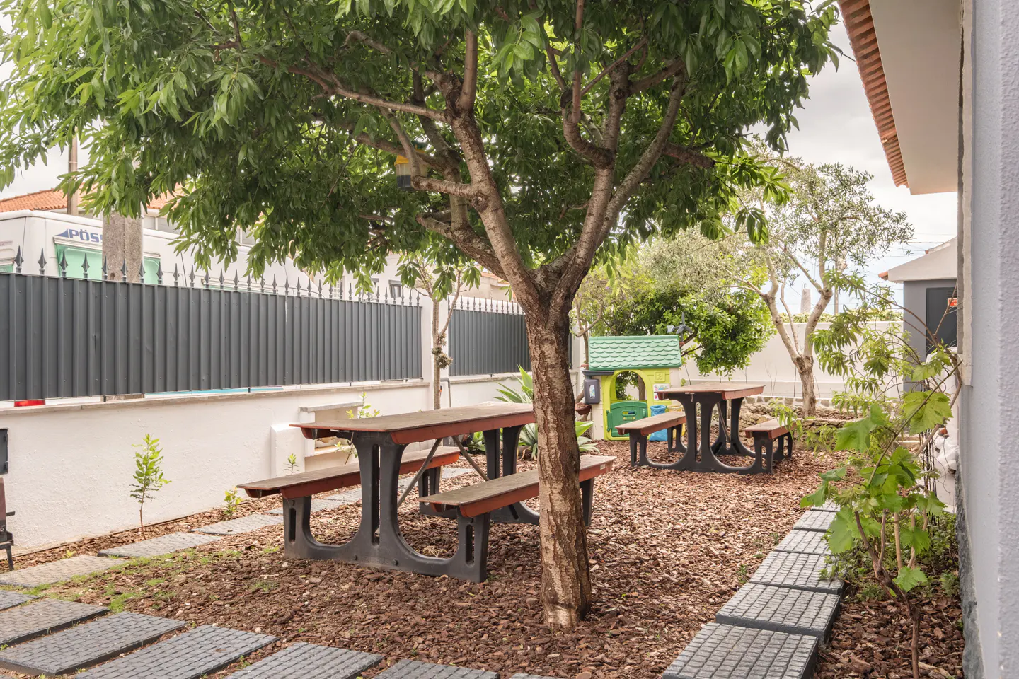 A backyard with two picnic tables under a tree, a green and yellow playhouse, and a gray fence in the background.