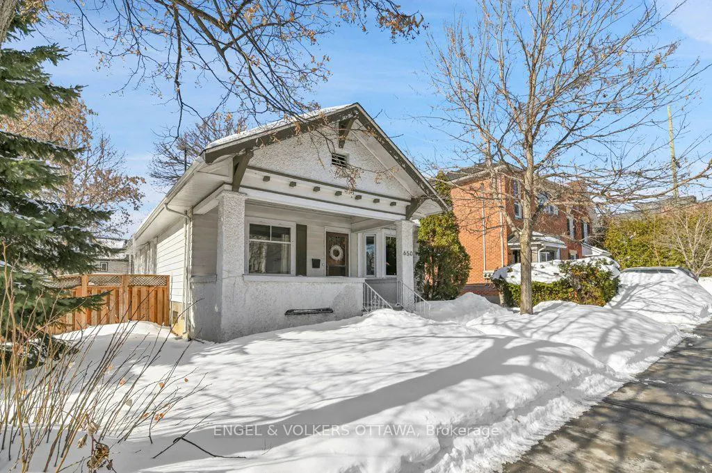 A white bungalow-style house with a front porch and snow-covered yard on a sunny winter day.