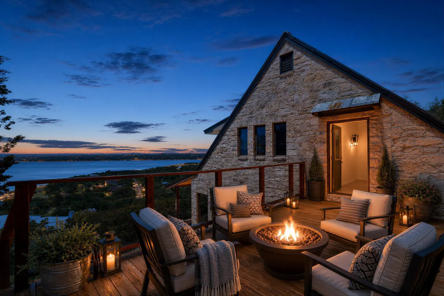 Outdoor deck with stone house, lake view, and fire pit. Chairs surround the fire pit, with lanterns and plants on the wooden deck.