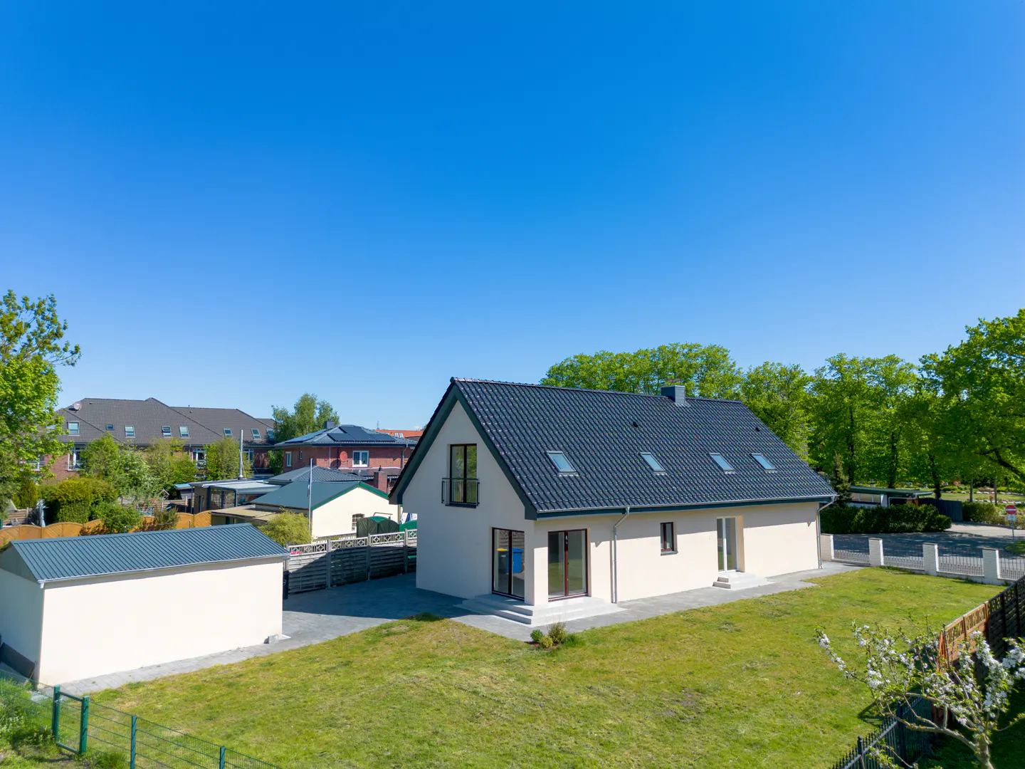 A modern, light-beige house with a dark gray roof sits on a green lawn under a clear blue sky. A matching shed is to the left.