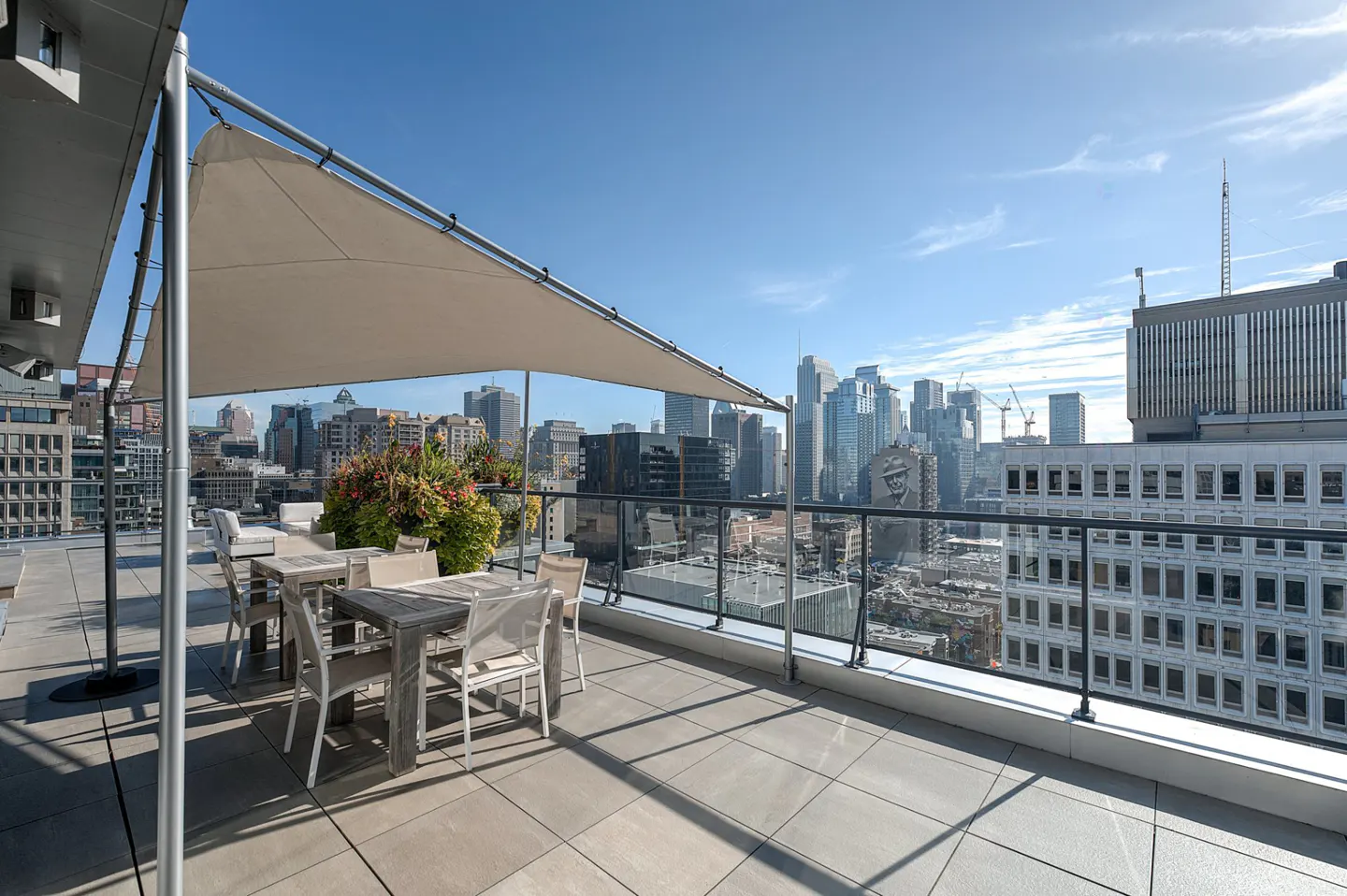 Rooftop terrace with dining set, shade sail, and potted plants overlooking a sunny city skyline with modern skyscrapers.