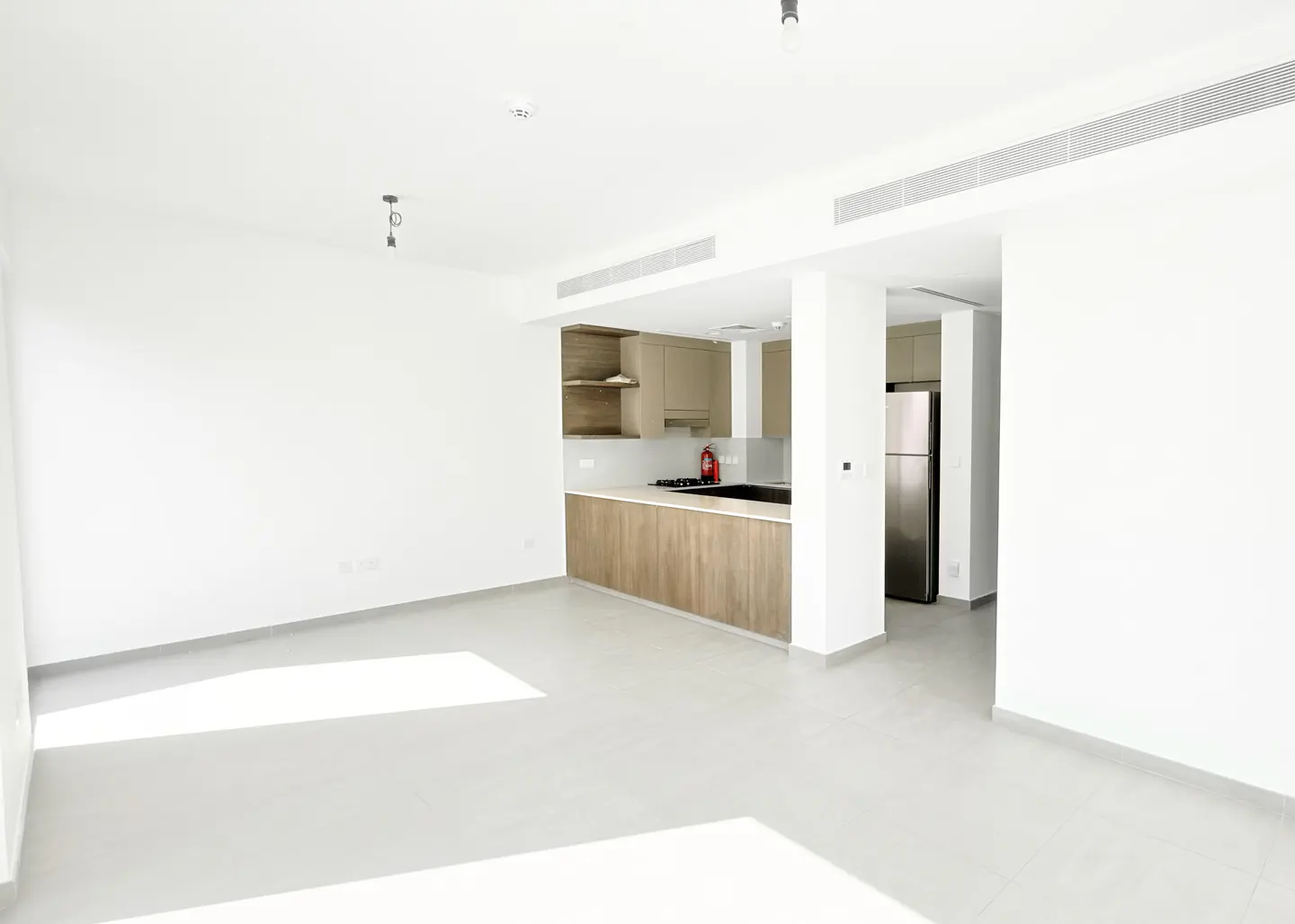 Bright, empty apartment with white walls and gray floors. An open kitchen features wood cabinets and stainless steel appliances.