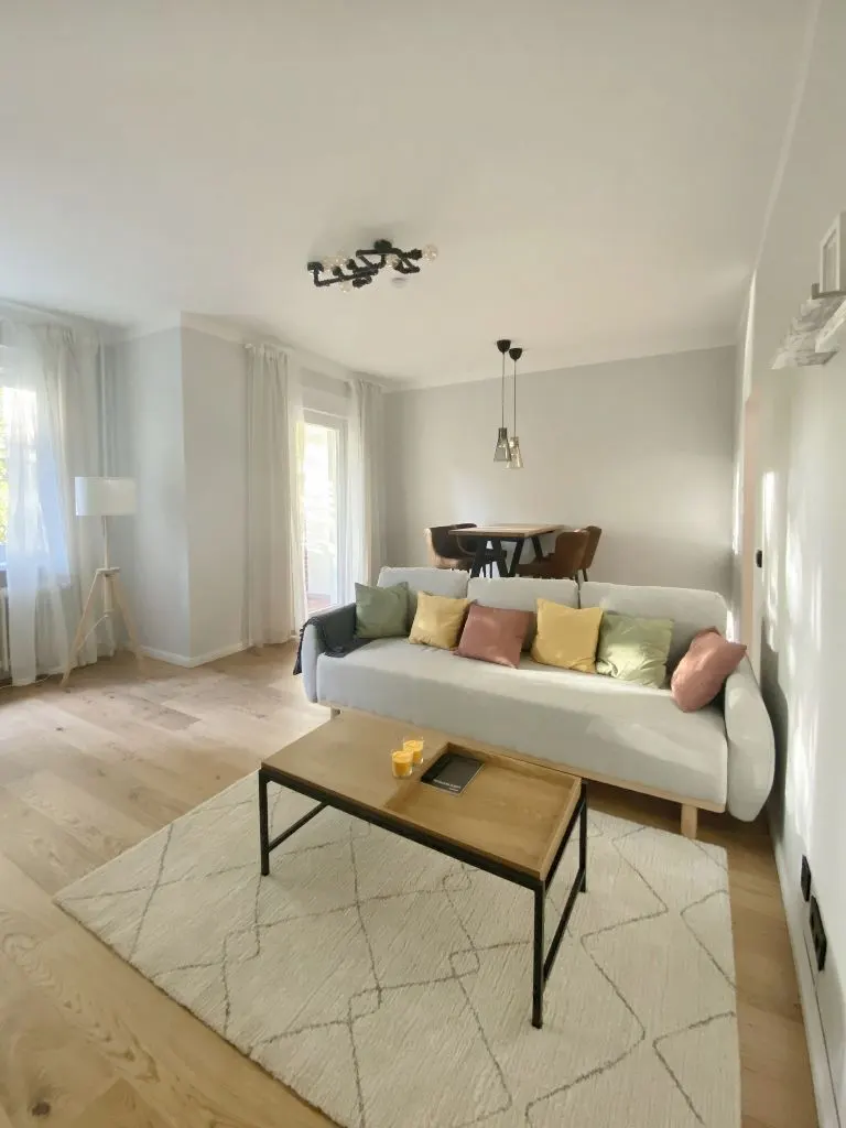 Bright living room with a light gray sofa, colorful pillows, and a wooden coffee table on a patterned rug. Dining table in the background.