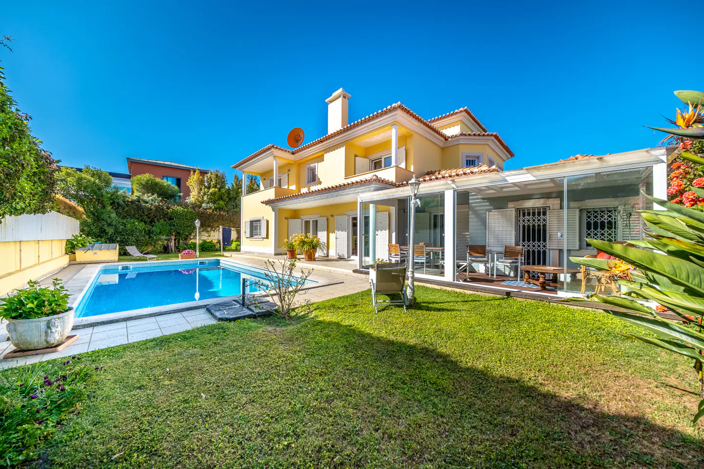 Two-story yellow house with a red tile roof, a pool, and a green lawn under a clear blue sky.