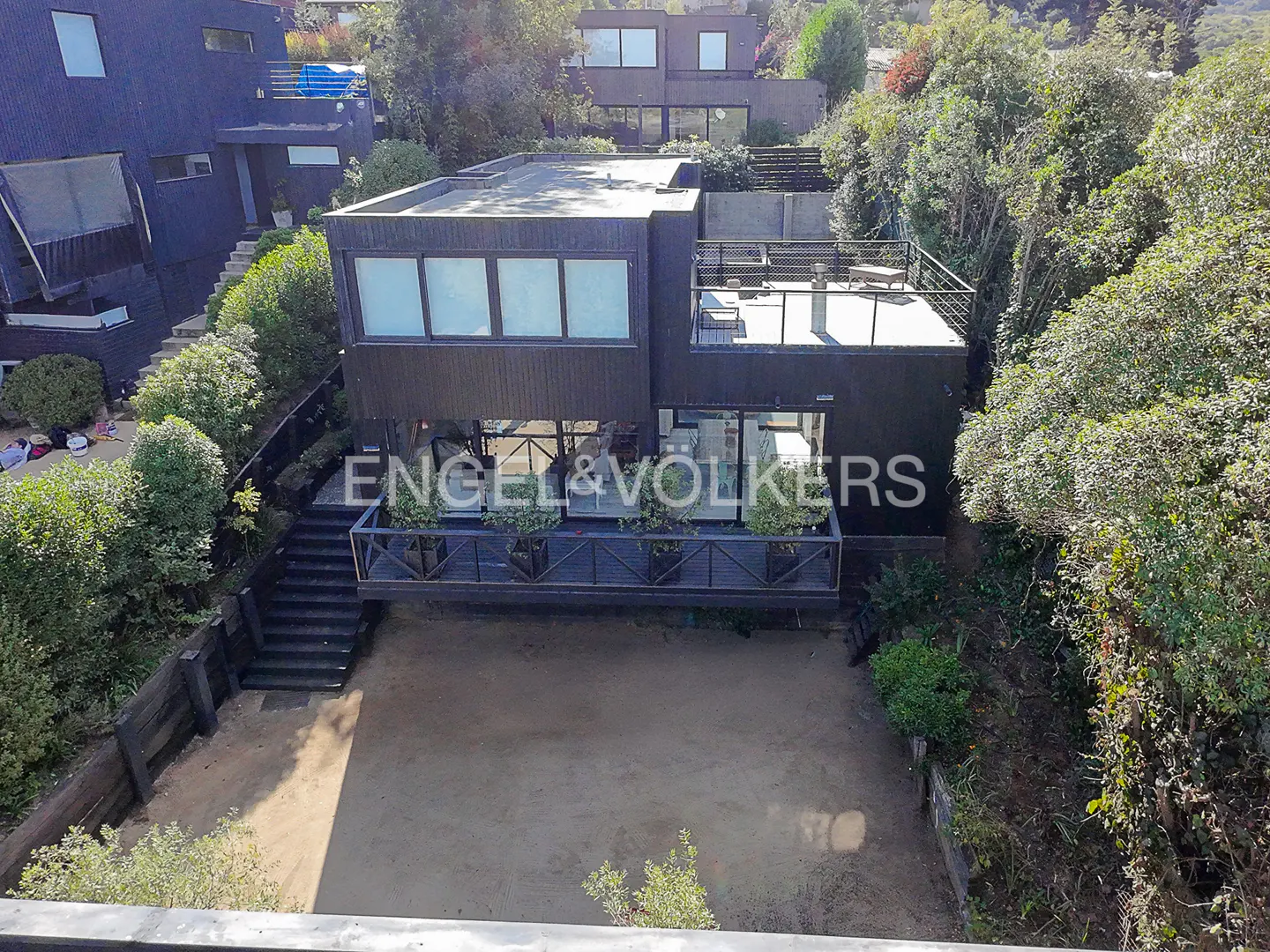 Aerial view of a modern, dark-colored house with a flat roof and a balcony, surrounded by lush greenery and other houses.