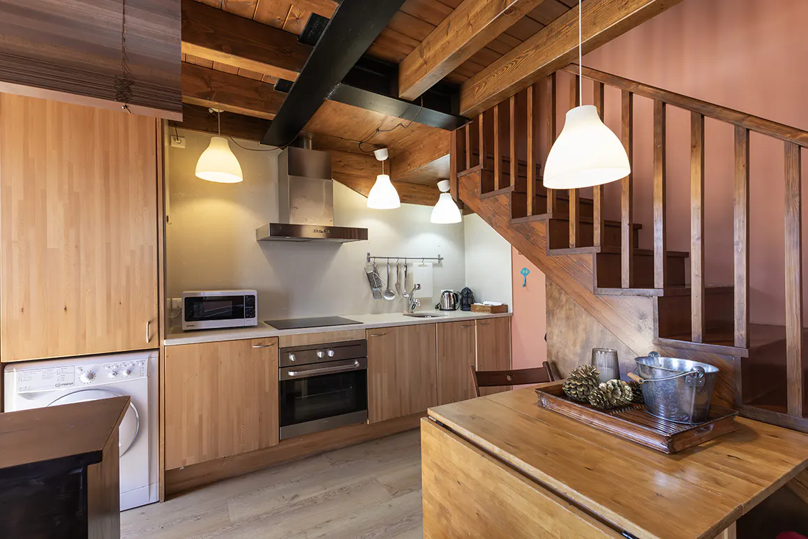 Cozy kitchen with wood cabinets, stainless steel appliances, and a wooden staircase. Pendant lights hang from the wood-beamed ceiling.