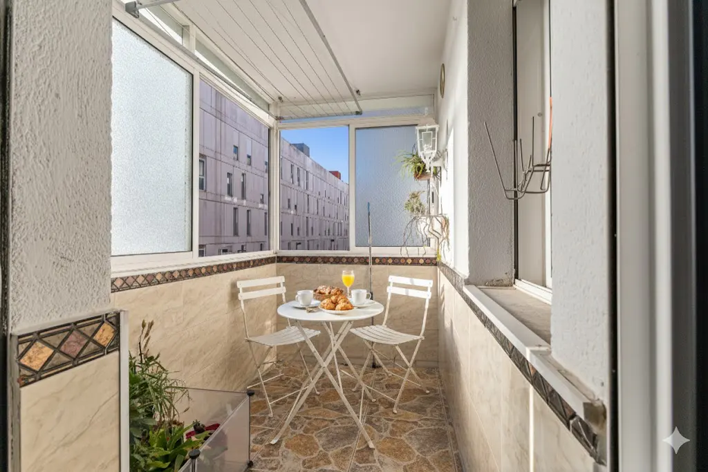 A bright balcony with a white table set for breakfast, including croissants, coffee, and orange juice. A building is visible through the window.